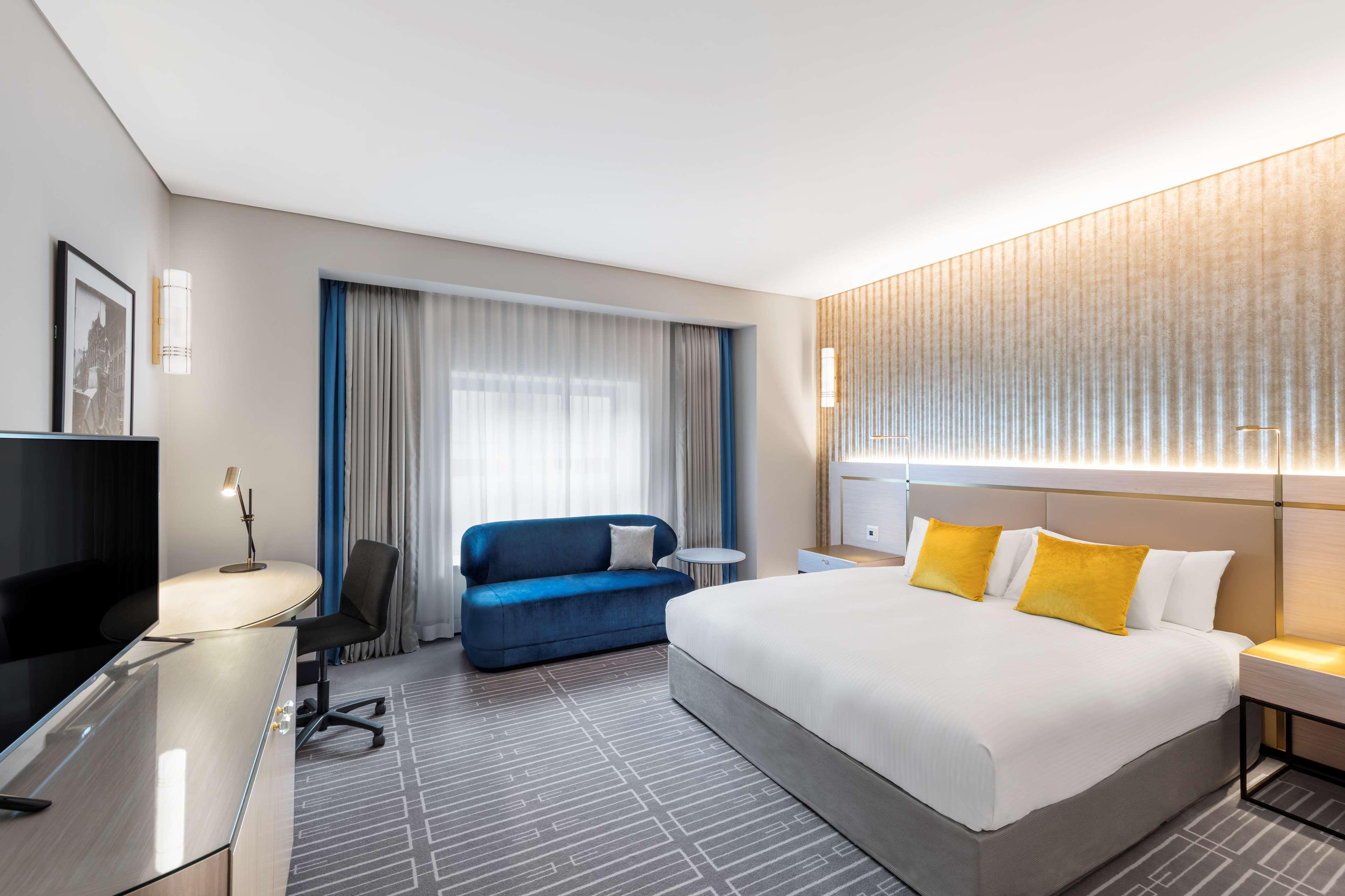 Radisson Blu Plaza Hotel Sydney Exterior photo The photo shows a modern hotel room featuring a spacious layout. It has a large bed with white linens and yellow accent pillows. Beside the bed, there is a bedside table and a contemporary lamp. There is a blue sofa and a coffee table near a set of l